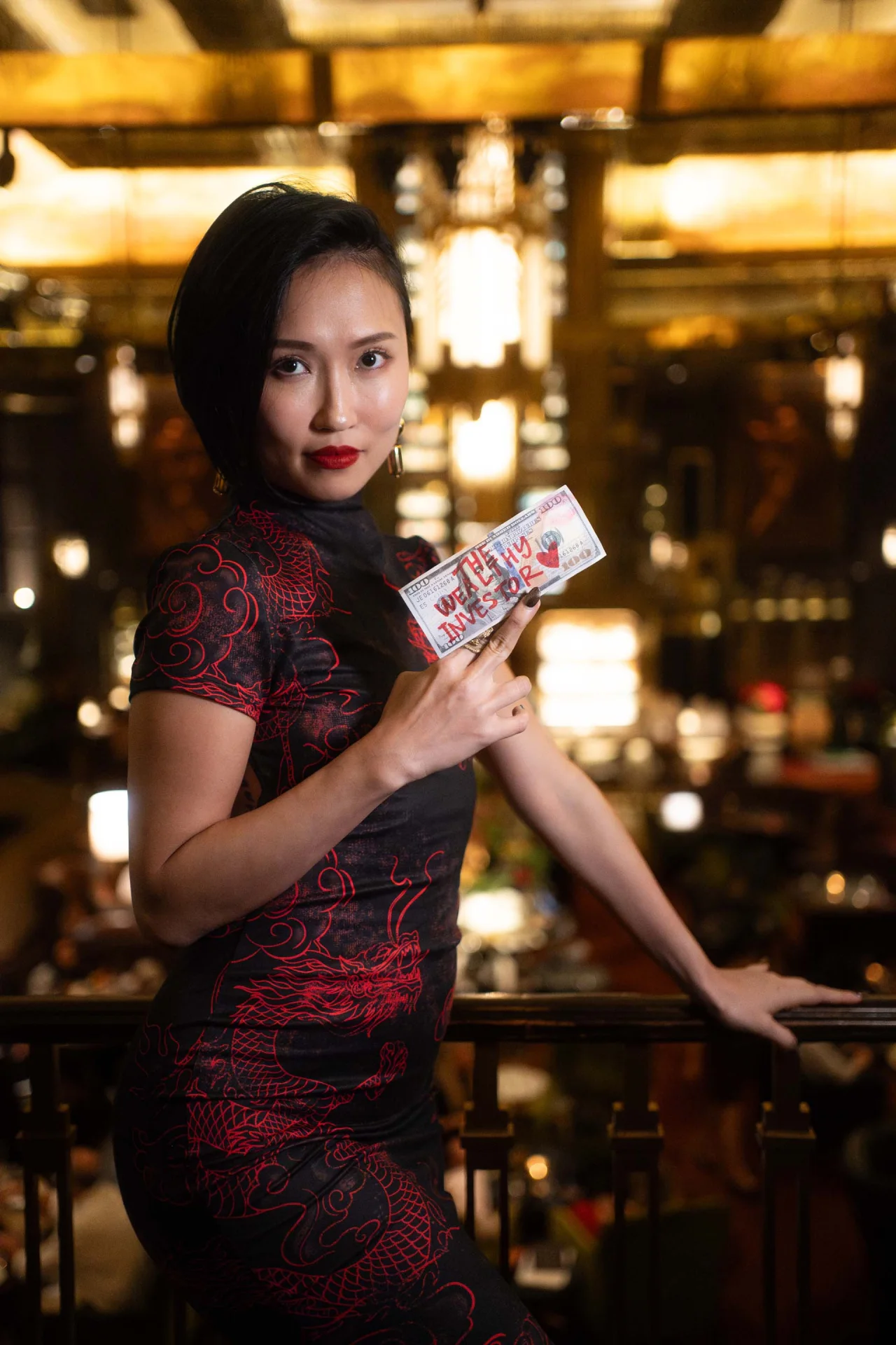 Crystal Chow at a bar in a red dragon qipao, holding a hundred-dollar note marked The Wealthy Investor