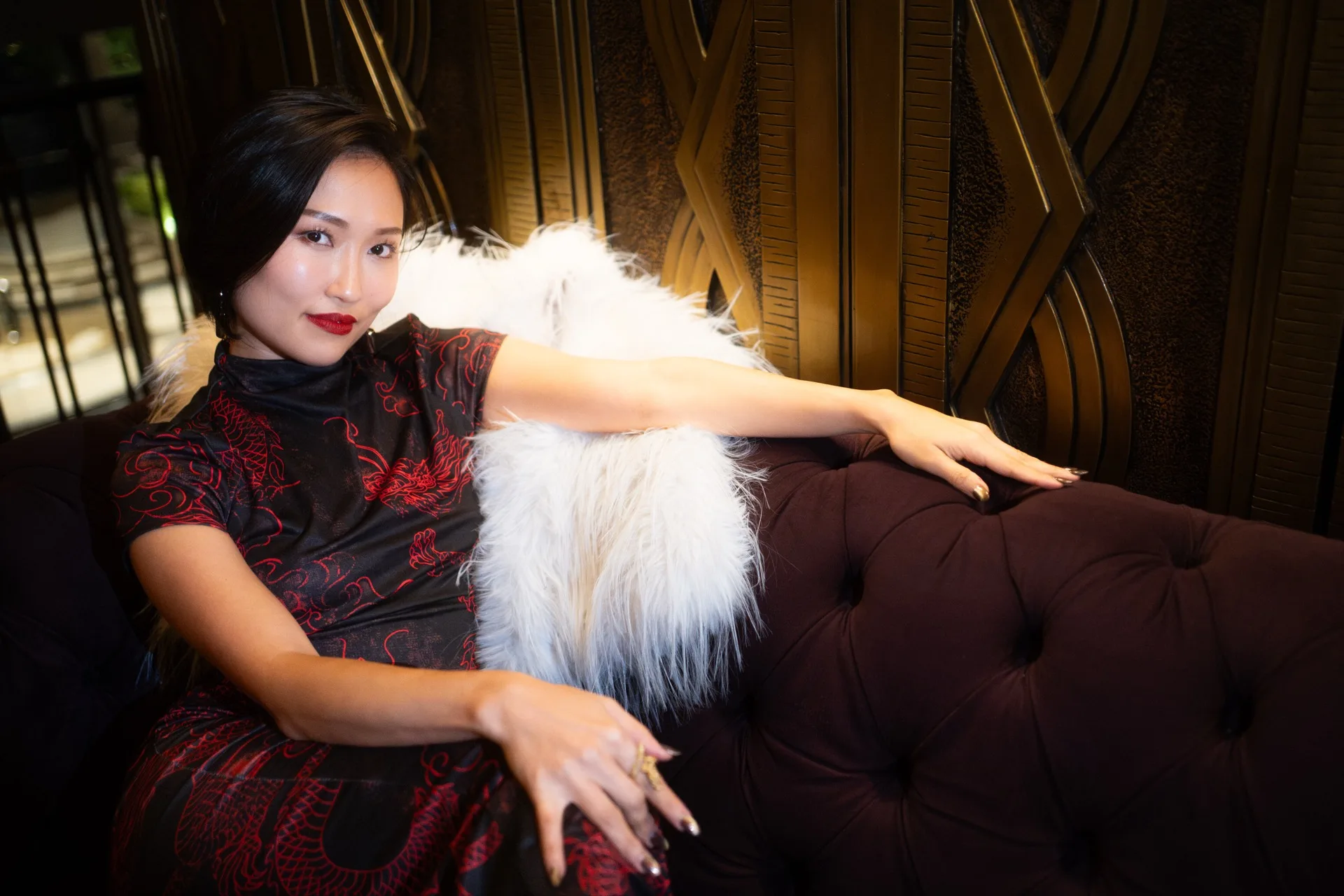 Crystal Chow reclining on a burgundy chesterfield in a red-and-black dragon qipao and white fur, against a gold art-deco screen