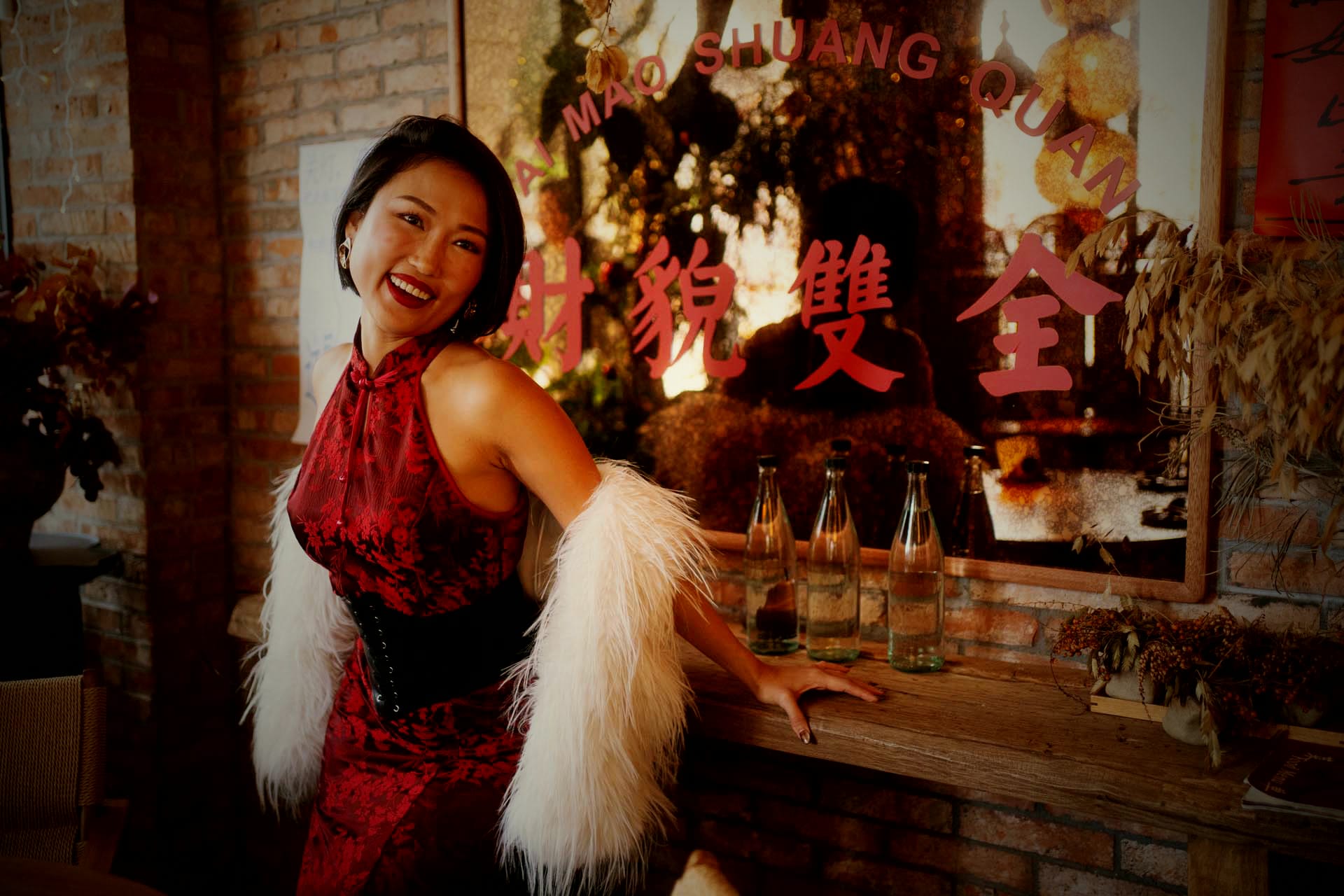 Crystal Chow laughing in a red lace cheongsam and feather stole, at a brick-walled bar