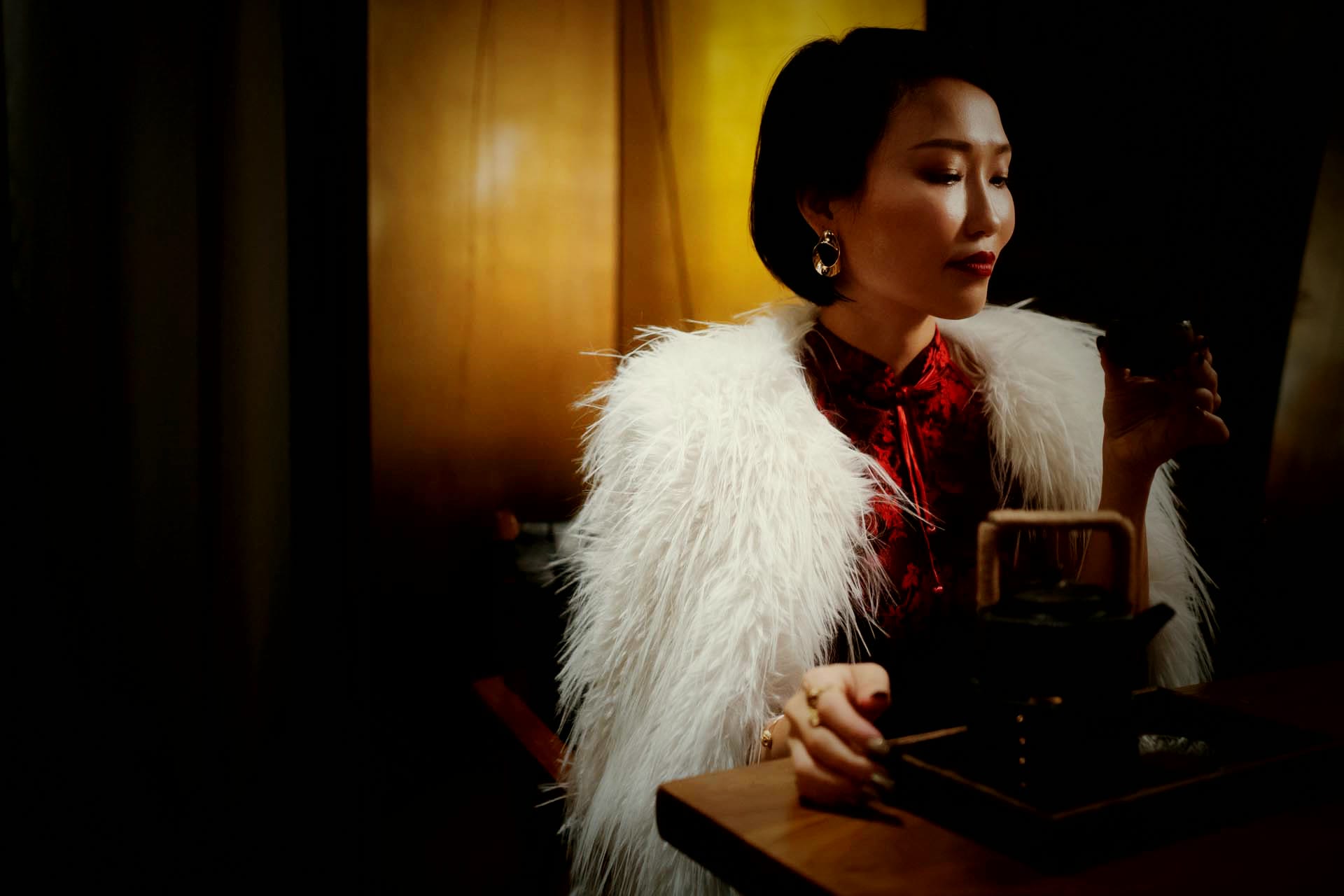 Crystal in profile at a dark tea table, red dress under a white feather stole