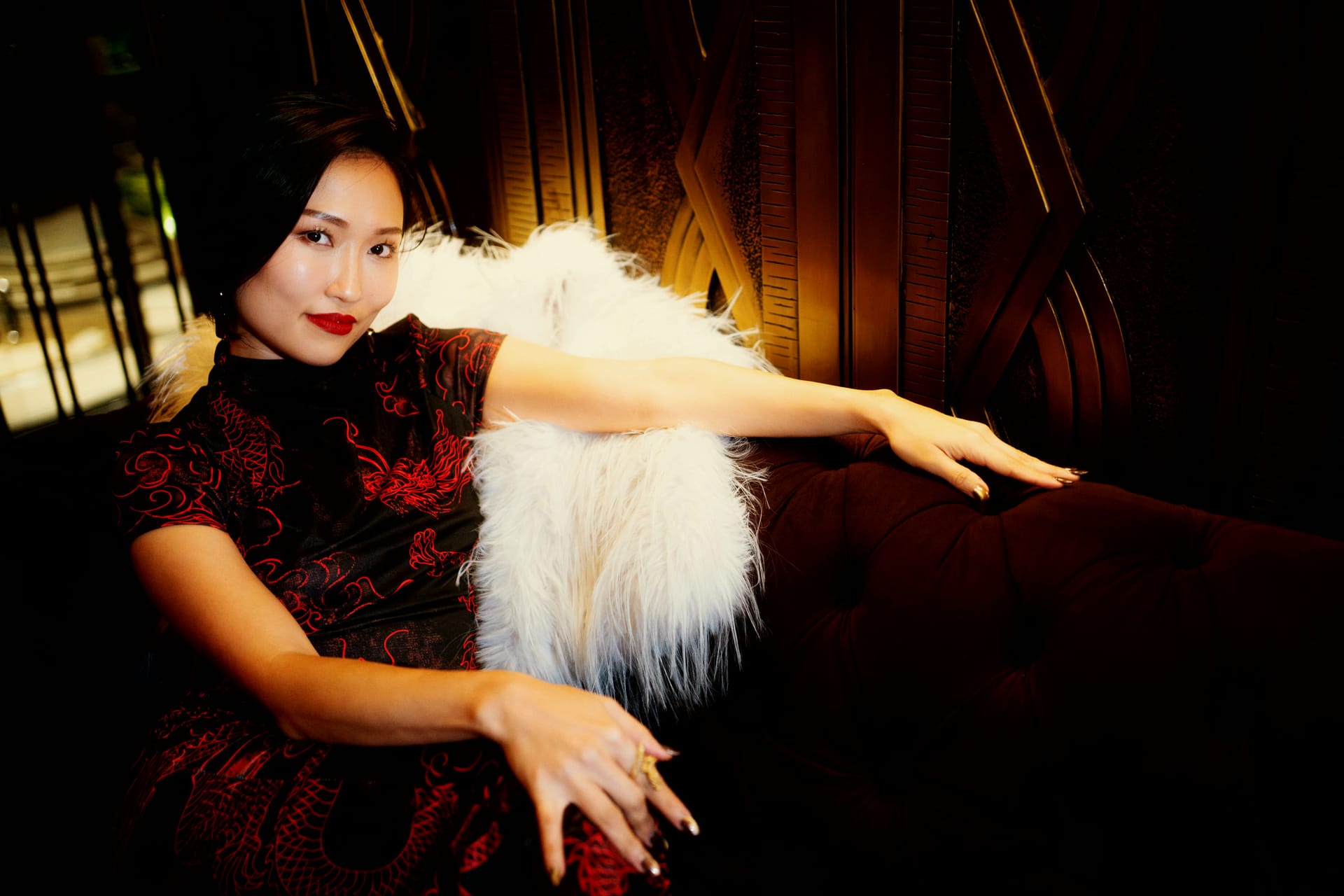 Crystal Chow in a red silk cheongsam and white feather stole, reclining in a gold-lit bar