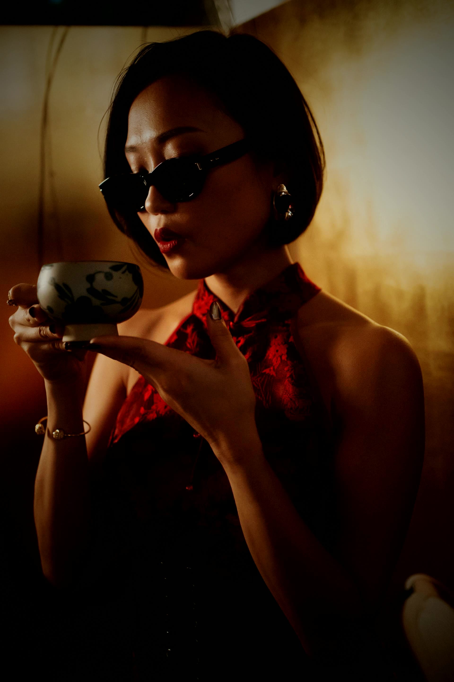 Crystal Chow in a red cheongsam and sunglasses, raising a painted teacup in gold lamplight