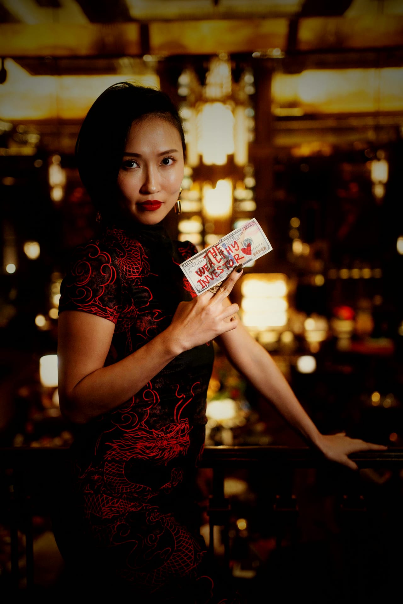 Crystal Chow at a dark bar in a black cheongsam with red dragon embroidery, holding a hand-lettered note